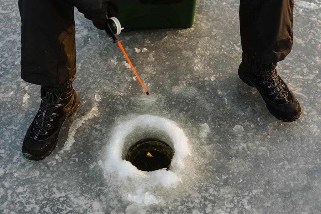 Person ice fishing on frozen lake surface with fishing rod and winter boots.